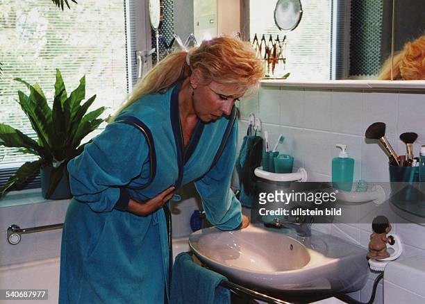 Eine Frau im Bademantel beugt sich im Badezimmer über das Waschbecken und hält sich die Hände auf den Bauch. Bauchschmerzen, Übelkeit,...