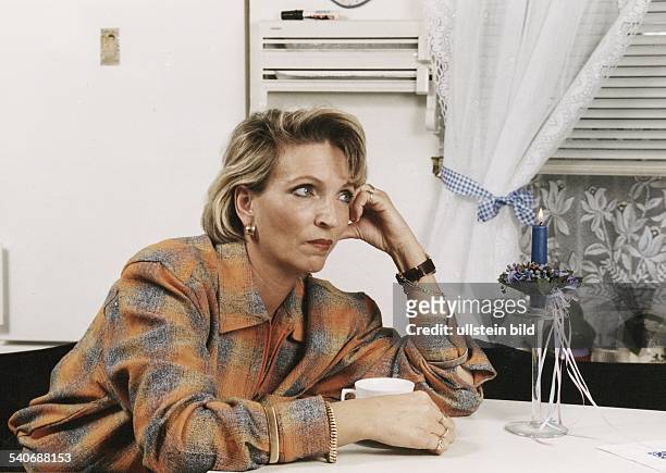 Eine Frau sitzt mit nachdenklichem Gesichtsausdruck und auf die Hand gestütztem Kopf am Tisch. Sie trägt Goldschmuck an Armen, Fingern und Ohren. Vor...
