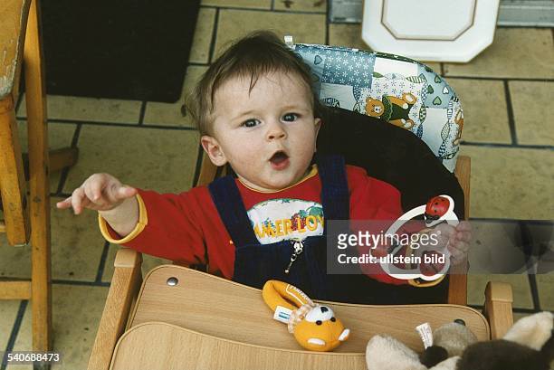 Ein siebenmonate altes Kleinkind sitzt auf einem Kinderstuhl mit integrierter Tischplatte und hat eine Rassel in der Hand. Undatiertes Foto.
