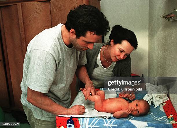 Familie: Junge Eltern mit ihrem Baby. Der Vater reinigt den Po des auf dem Wickeltisch liegenden Säuglings, während die Mutter dabei zusieht und sich...