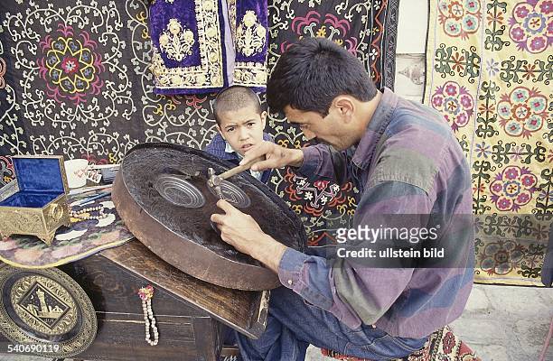 In seiner kunstgewerblichen Werkstatt in Buchara, Usbekistan, meißelt ein Mann Ornamente in ein rundes Gefäß. Ein kleiner Junge schaut ihm dabei zu....