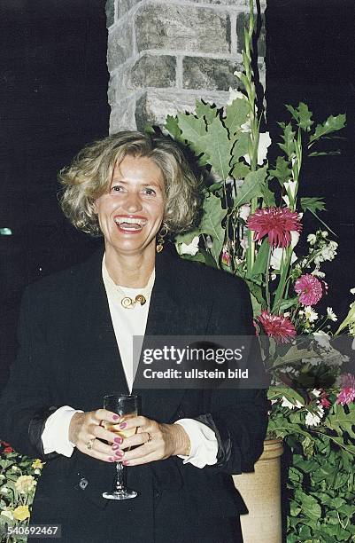 Die Kinderbuchautorin Brigitte Blobel steht mit einem Sektglas in beiden Händen und lacht herzlich. Im Hintergrund Blumengestecke. Aufgenommen...