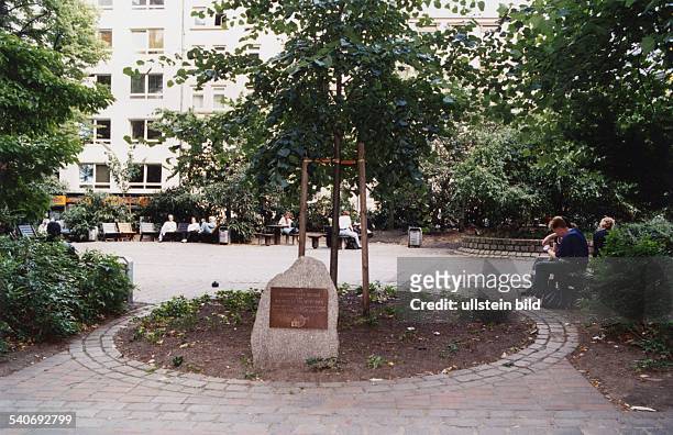 Der Gertrudenkirchhof mit einem Gedenkstein. Rund um den Platz stehen unter Bäumen Stühle, die Passanten zum Verweilen einladen. .