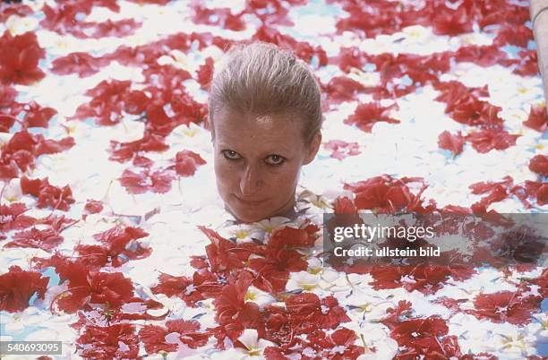 Eine Frau badet in einem Pool, an dessen Wasseroberfläche die Blüten von Aralien und Hibiskus schwimmen. Bad; Ayurveda-Kur; Frauenkopf. Undatiertes...