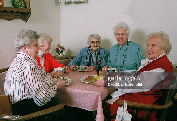 Eine Gruppe fröhlicher alter Frauen trifft sich am Mittagstisch zum Essen. Die Damen löffeln Suppe aus Suppentassen und als Hauptgericht gibt es...
