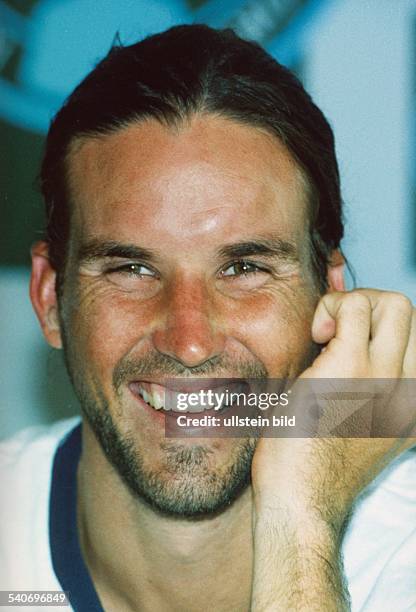 Der australische Tennisspieler Patrick Rafter lacht fröhlich in die Kamera. Er trägt einen Drei-Tage-Bart und hält die linke Hand an seine Wange....