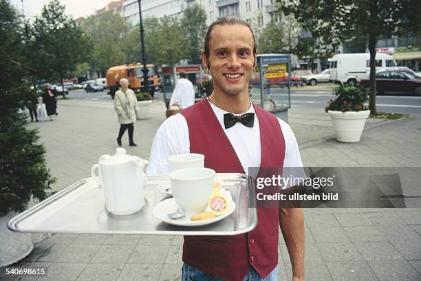 Auf dem Tablett des Kellners stehen zwei große Kaffeetassen und eine Kaffeekanne. Der Kellner steht auf einem breiten Bürgersteig und trägt zu Jeans...