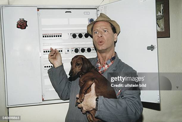 Der Kabarettist Tom Gerhardt vor einem Sicherungskasten als 'Hausmeister Krause' mit Hut und dem Dackel 'Bodo' auf dem Arm. Undatiertes Foto.