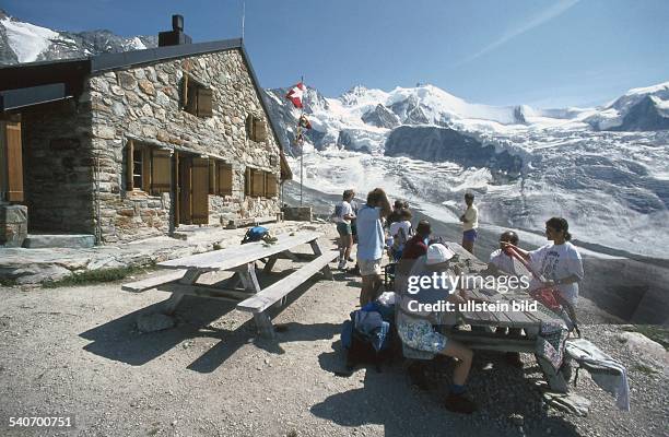 Wanderer halten Brotzeit an Holztischen- und Bänken vor der Cabane d' Ar Pitetta bei Zinal in den Walliser Alpen. Im Hintergrund das Zinalrothorn und...