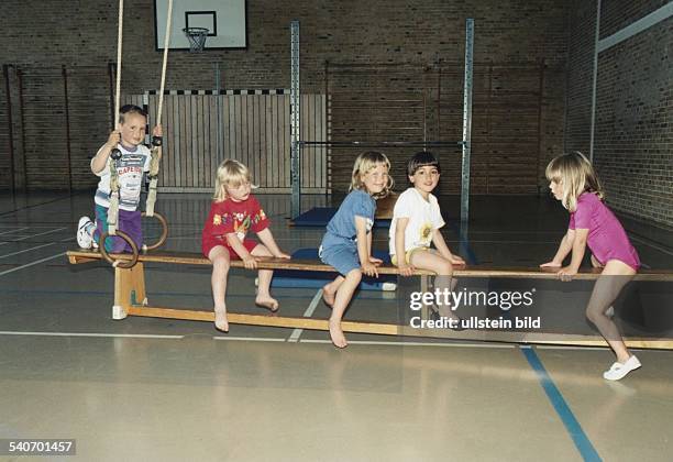 Jungen und Mädchen in Sportkleidung sitzen beim Turnunterricht auf einer Bank in einer Sporthalle. Ein Kind hält sich an Ringen fest....