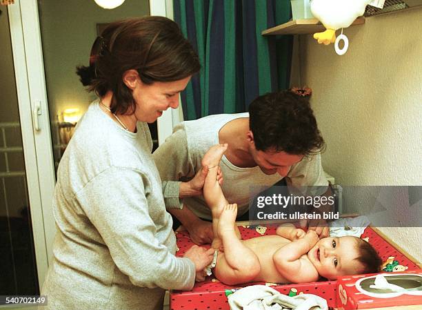 Familie: Eine Mutter wischt ihrem auf dem Wickeltisch liegenden Säugling den Po, während der Vater sich zu seinem Sohn herunterbeugt. .