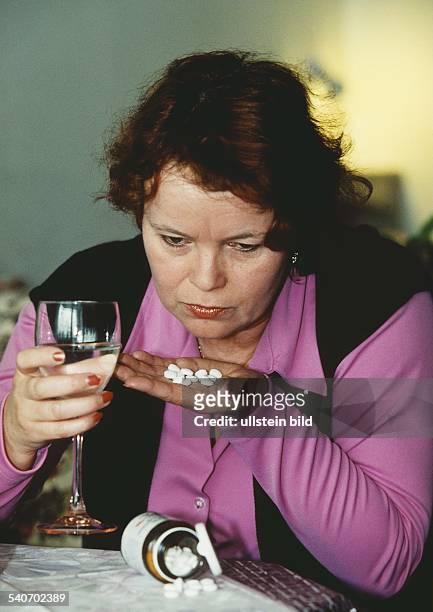 Suchtgefahr: eine Frau sitzt an einem Tisch, in der einen Hand mehrere Tabletten, in der anderen ein vollgeschenktes Weinglas. Alkoholmissbrauch;...