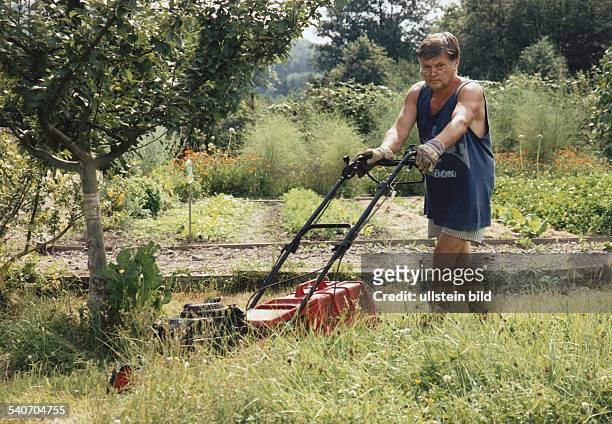 Mann in Shorts und T-Shirt mit Rasenmäher im Garten. Undatiertes Foto.