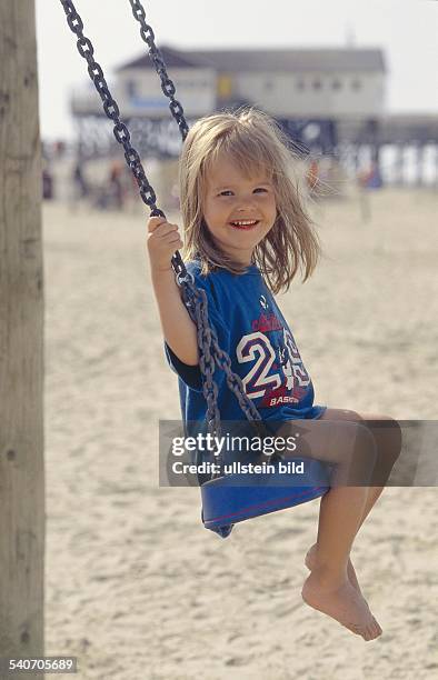 Ein kleines Mädchen sitzt lächelnd auf einer Schaukel am Strand. Undatiertes Foto.