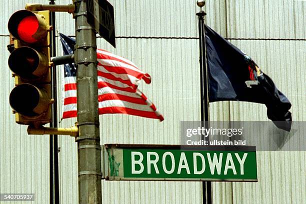 Der Broadway in New York: Das Straßenschild neben einer Ampelanlage, die Ampel zeigt rot an. Dahinter weht dieamerikanische Flagge, neben einer...