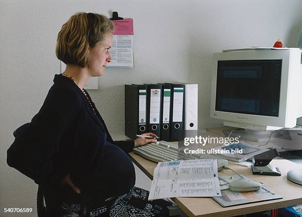 Eine schwangere Frau hält sich am Computerarbeitsplatz den Rücken. Auf ihrem Schreibtisch stehen Aktenordner, sie blickt konzentriert auf den...