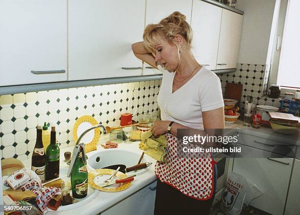 Eine Hausfrau mit Schürze und Putzlappen stützt sich erschöpft gegen den Einbauschrank ihrer Küche und betrachtet das abzuwaschende Geschirr sowie...