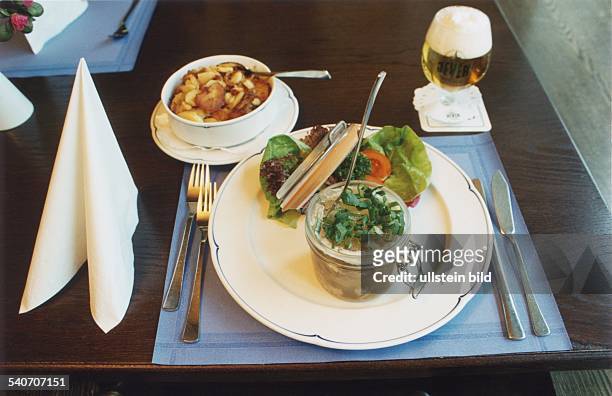 Auf einem mit drapierter Stoffserviette, Set und Besteck gedeckten Restauranttisch: ein geöffnetes Weckglas mit Sauerfleisch in Aspik, serviert auf...