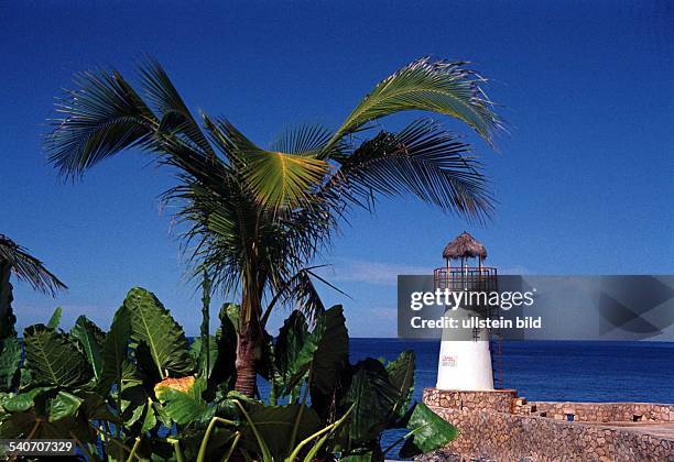 Kleiner Leuchtturm an der von Palmen und anderen exotischen Grünpflanzen gesäumten Felsenküste von Negril auf Jamaica. .