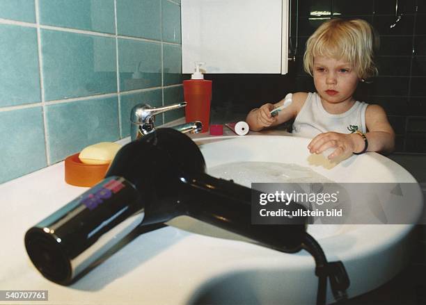 Ein Kind hält über einem mit Wasser gefüllten Waschbecken seine Zahnbürste. Auf der gegüberliegenden Seite des Waschbeckens liegt ein elektrischer...