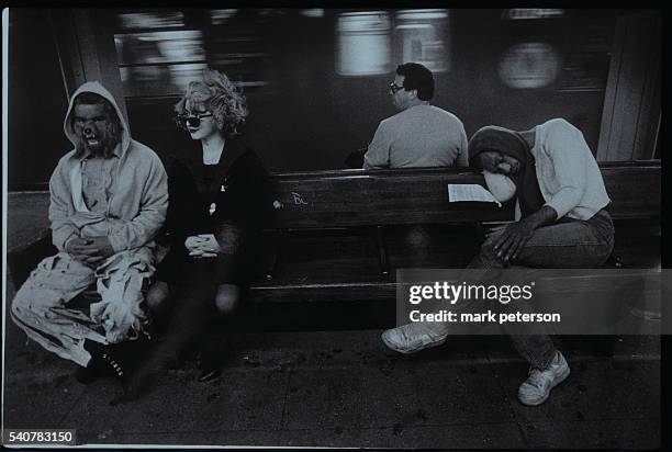 Homeless man sleeps next to two people wearing costumes while waiting for the subway.