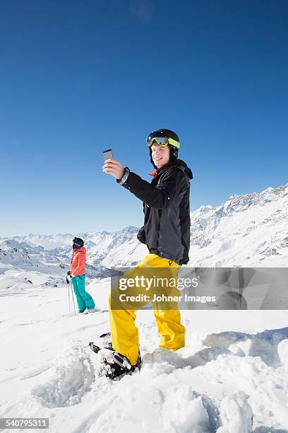 young man wearing skiwear taking picture with cell phone - sunny winter stock pictures, royalty-free photos & images