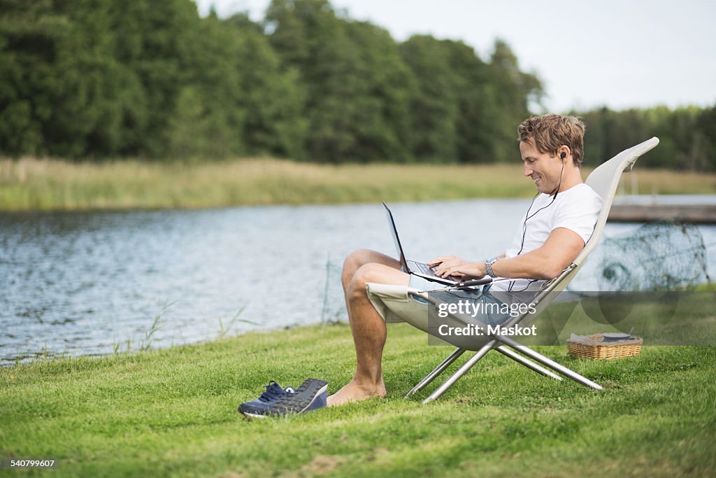 Full length side view of mature man using laptop on deck chair at lakeshore