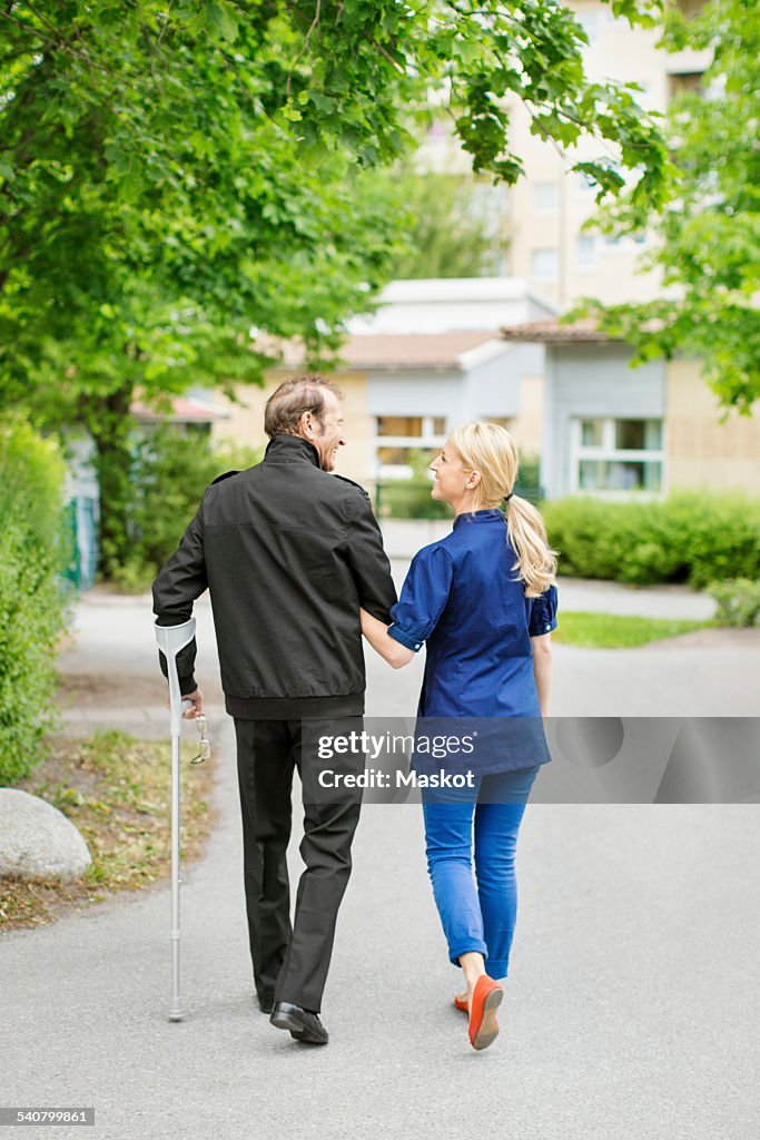 Full length rear view of female caretaker walking with disabled senior man on street