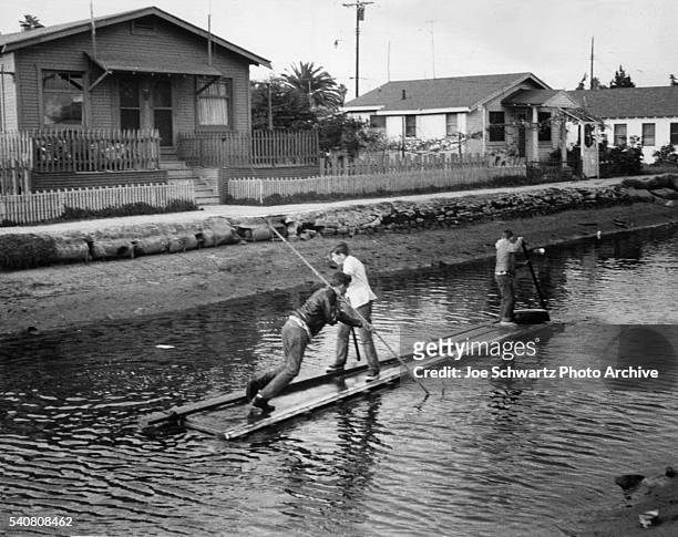 Three boys travel a canal in Venice, California, by punting a makeshift raft.