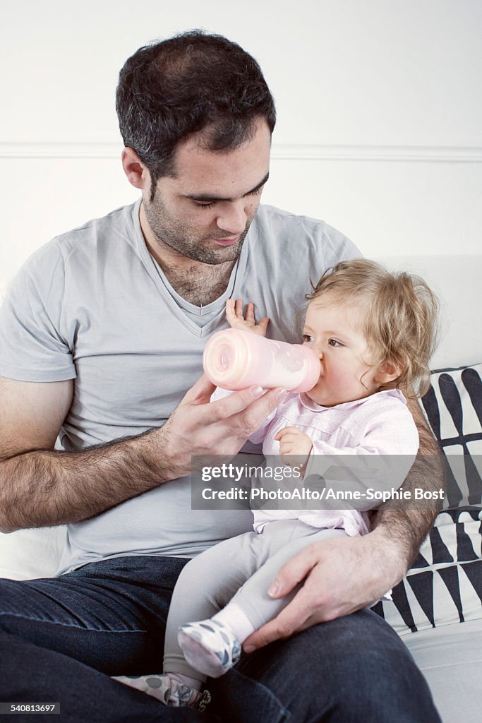 Father holding baby girl on lap, feeding her with bottle