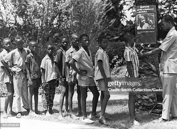 The Smallpox Eradication and Measles Control Programs, West Africa, 1968. Vaccination of boys in West Africa while standing in front of a poster...