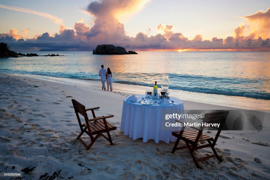 Romantic couple walking at sunset on beach with dinner table in foreground.Cousine Island.Seychelles