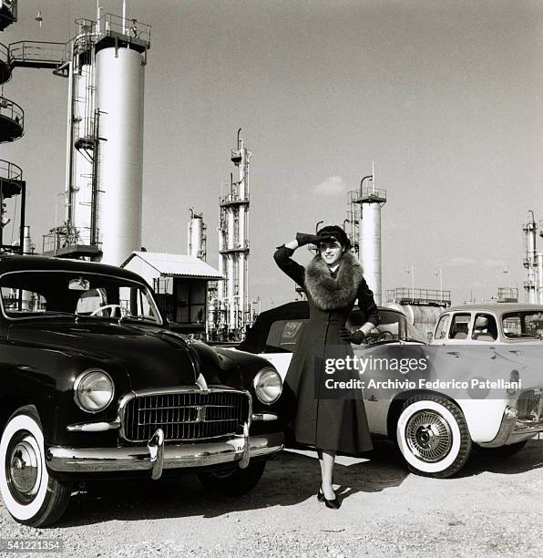 Woman wearing a fitted coat and fur stole poses between a Fiat 1400 saloon car and a sporty Fiat 1100.