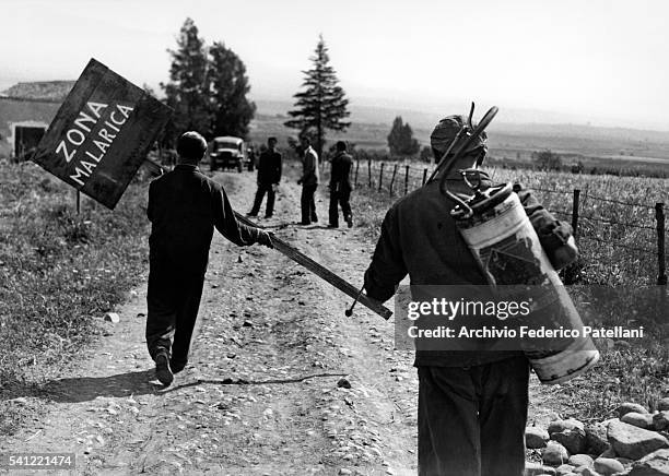 Two men bearing a fumigation tank and a makeshift sign make their way along a country track.