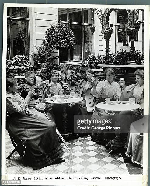 GOURP OF WOMEN SITTING AT TABLES WITH GLASSES RAISED IN A GARDEN CAFE IN BERLIN, GERMANY. PHOTOGRAPH, TURN OF THE CENTURY.