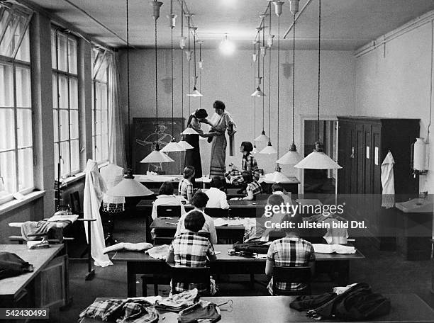 Textil- und Modeschule der Stadt Berlin:Blick in einen Unterrichtssaal. EinKleidungsstück wird gesteckt,Schülerinnen sehen zu.- 1933