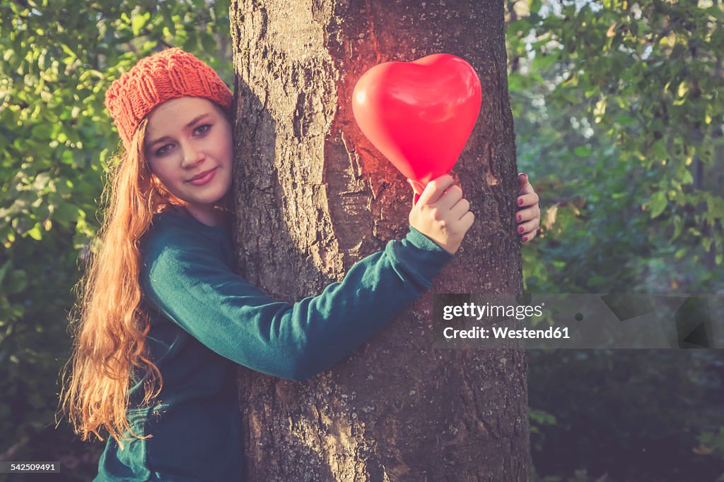 Portrait of smiling teenage girl with heart shaped balloon hugging a tree