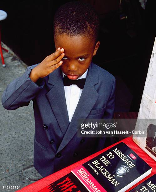 Young Muslim boy in a suit salutes, while standing next to a display of books, one of which is my Elajiah Muhammad, one of the founders of the Nation...