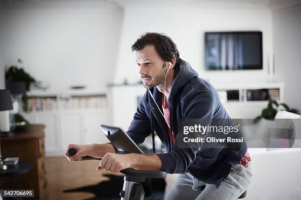 man with earbuds on exercise bike at home - exercise bike fotografías e imágenes de stock