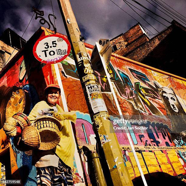 Colombian street vendor walks around a street corner, covered in colorful graffiti artwork and posters, in La Candelaria, Bogota, Colombia, 19...