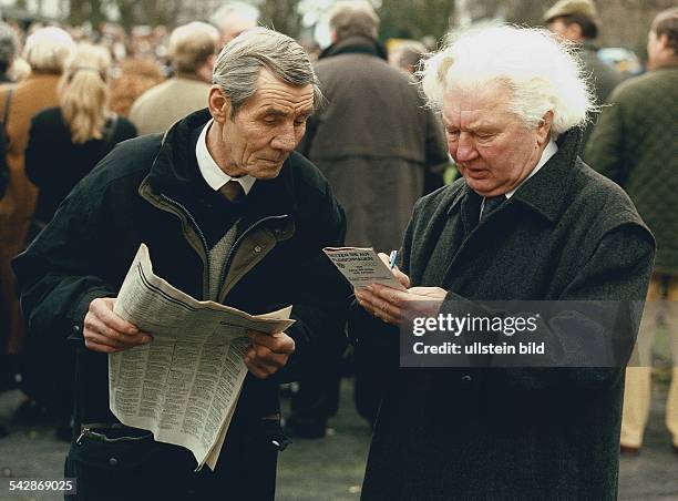 Zwei alte Männer beschäftigen sich mit den Quoten des Galopprennens. Einer von ihnen hält eine Zeitung in der Hand und sieht dem anderen Mann dabei...