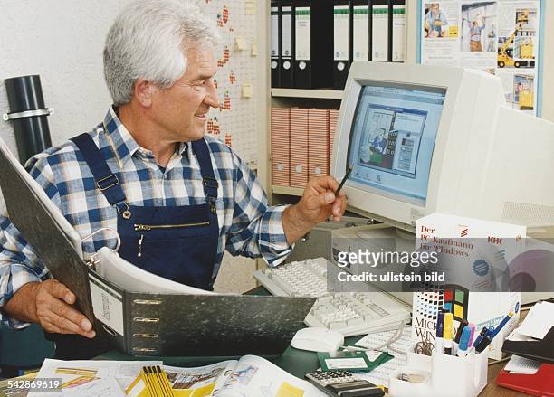 Ein Handwerker sitzt an einem vollen Schreibtisch. In der rechten Hand hält er einen Aktenordner, in der linken einen Bleistift, mit dem er auf den...
