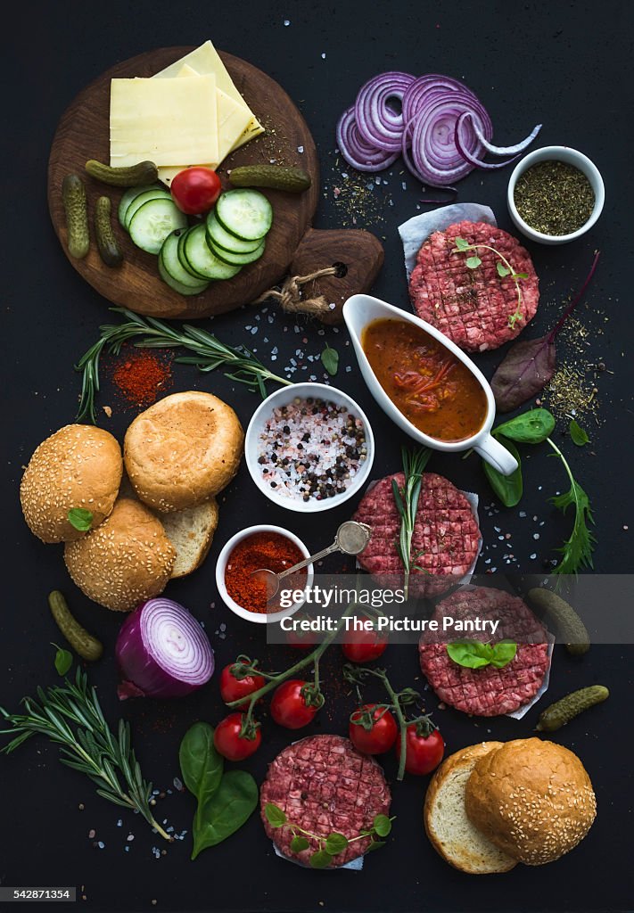 Ingredients for cooking burgers. Raw ground beef meat cutlets, buns, red onion, cherry tomatoes, greens, pickles, tomato sauce, cheese, herbs and spices over black background, top view.