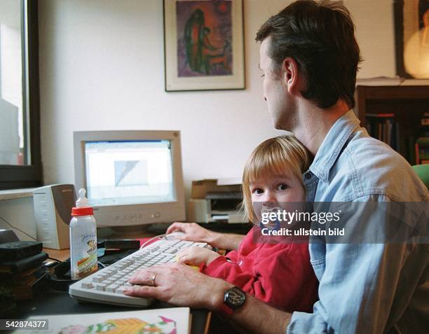 Ein Mann sitzt in häuslicher Umgebung mit einem kleinen Kind auf dem Schoß an seinem Computerarbeitsplatz. Das Kind hat einen Schnuller im Mund, sein...