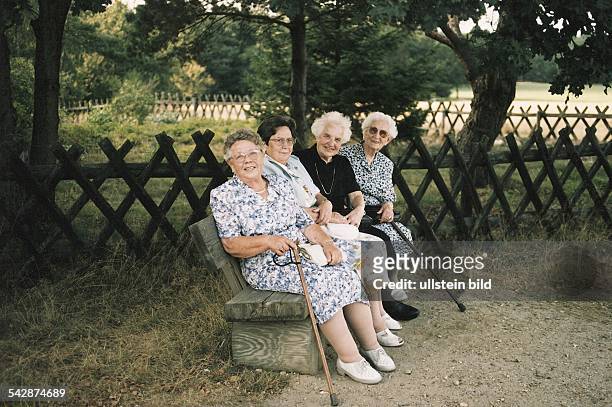 Rentnerinnen treffen sich auf einer Parkbank, die unter Schatten spendenden Bäumen aufgestellt ist. Zwei der Seniorinnen haben ihre Gehstöcke dabei....