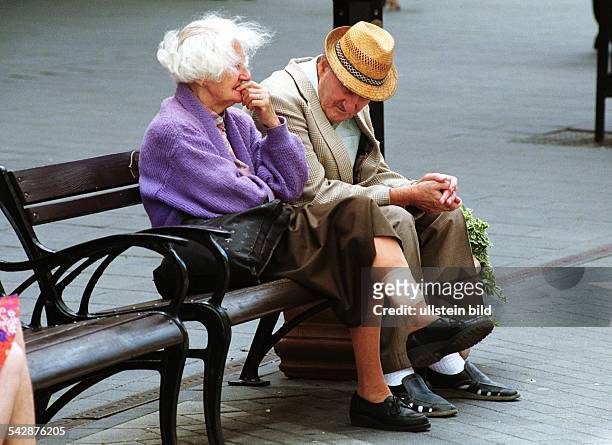 Ein altes Paar ruht sich auf der Straße auf einer Bank aus. Eng nebeneinander sitzend hält der Mann seinen mit einem Strohhut bedeckten Kopf gesenkt,...