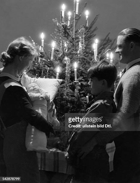 Familie - Vater, Mutter mit Baby auf dem Arm und Sohn,stehen vor dem Weihnachtsbaummit brennenden Kerzen.- 1938Foto: Max Ehlert