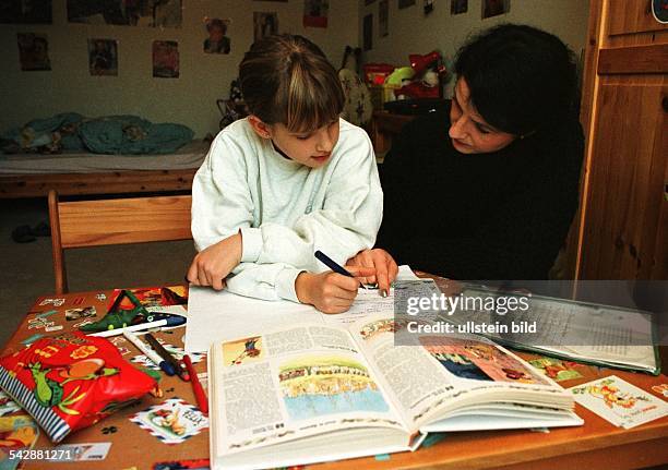 Mutter und Tochter sitzen im Kinderzimmer an einem Schreibtisch bei den Hausaufgaben. Der unaufgeräumte Schreibtisch ist mit Stiften und Kleinkram...