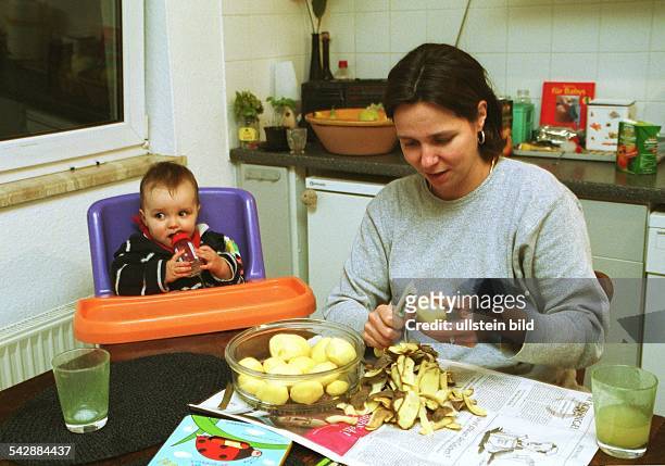 Eine junge Frau schält am Küchentisch Kartoffeln während ihr Baby im Kinderhochstuhl sitzt und an seiner Nuckelflasche saugt. Haushalt und Kind. .