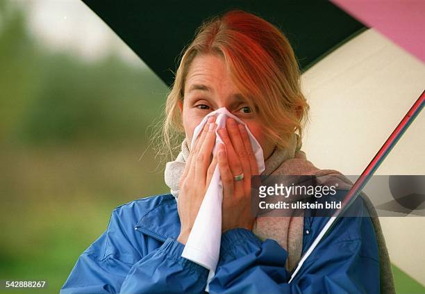 Eine Frau mit Schal unter einem Regenschirm schnäuzt in ein Taschentuch. Schnupfen; Erkältung; Grippe; Erkältungswetter. Symbolbild .